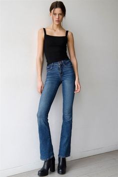 Woman in a black tank top and blue jeans leaning against a white wall