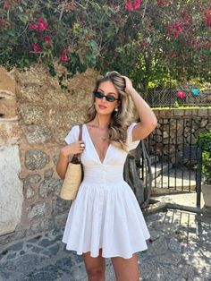 Woman in a white dress and sunglasses posing outdoors by a stone wall