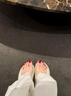 Feet with red nail polish wearing white slippers and pants on a dark carpet