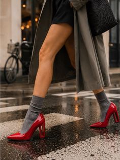 Person in a trench coat, red heels, and grey socks walking on a wet street