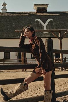 Woman in a brown jacket and cowboy boots leaning on a wooden fence at a ranch