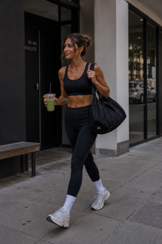 Woman in black athletic wear walking outdoors with a drink and a gym bag