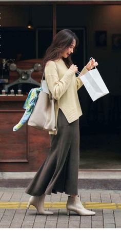 Woman in a beige sweater and long skirt walking on a street with a shopping bag