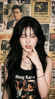 Woman with long black hair winking, wearing a tank top and jewelry, against a poster wall