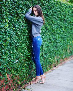 Woman in blue jeans and a gray sweater leaning against an ivy-covered wall