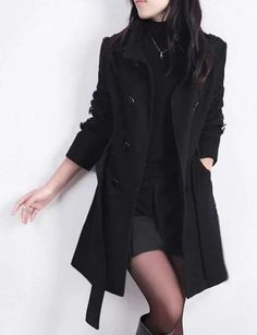 Woman in a black coat, turtleneck, shorts, and tights against a white wall