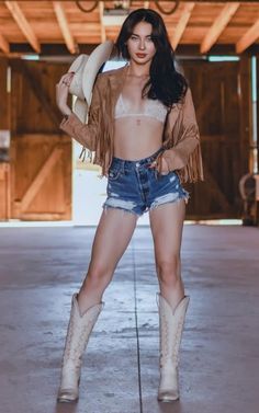 Woman in a fringe jacket, denim shorts, and cowboy boots holding a cowboy hat