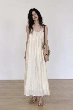 Woman in a long cream dress and sandals holding a straw bag indoors