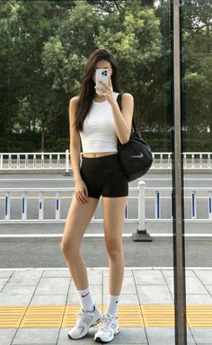 Woman in a white crop top and black shorts taking a mirror selfie outdoors