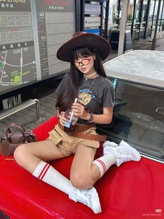 Woman in a cowboy hat and khaki shorts sitting on a red car outdoors