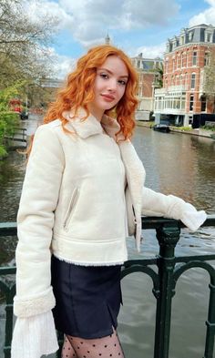 Woman with red curly hair in a white jacket and black skirt by a canal