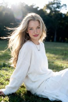 Woman with long blonde hair and freckles sitting on grass in a white sweater