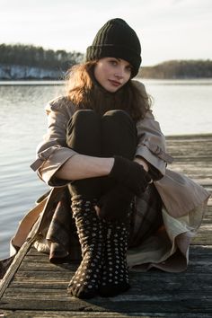 Woman in a black beanie and trench coat sitting on a wooden dock by a lake