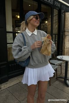 Woman in a baseball cap and pleated skirt holding coffee and a pastry outdoors