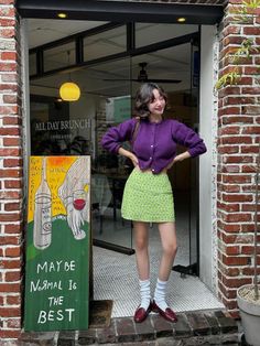 Woman in a purple cardigan and green mini skirt posing outside a cafe