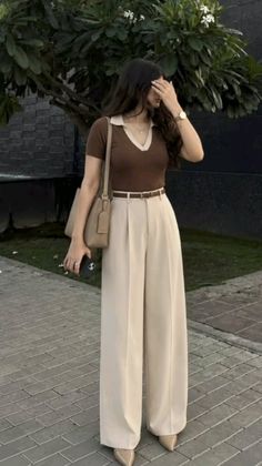 Woman in a brown polo shirt and wide leg pants holding a phone outdoors