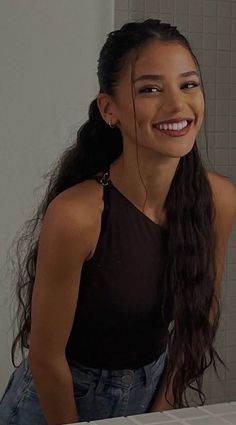 Woman with long dark hair and a one-shoulder top smiling indoors