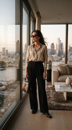 Woman in a blouse and wide leg pants looking out at a cityscape from indoors