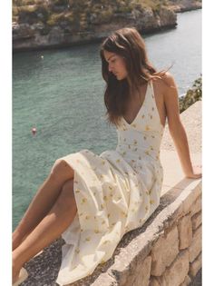 Woman in a white summer dress with lemon print sitting on a stone wall by the ocean