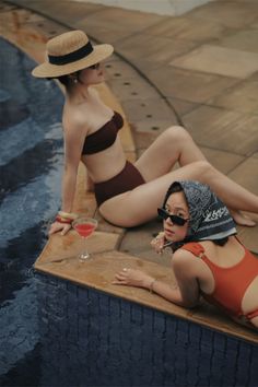 Two women in swimwear relaxing by a pool with a cocktail