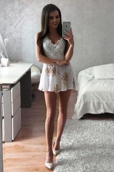 Woman in a white floral dress and high heels taking a mirror selfie in a bedroom