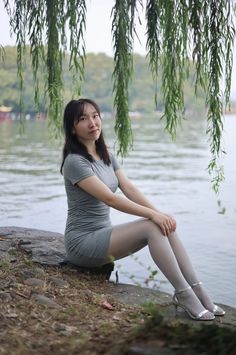 Woman in a grey dress and tights sitting by a lake under a willow tree