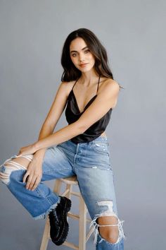 Woman with long dark hair wearing a black top and ripped jeans, sitting on a stool