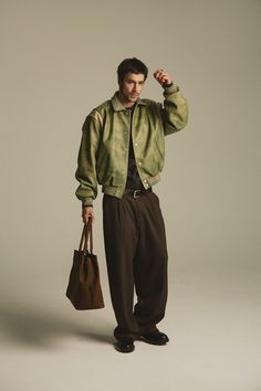 Man in a green bomber jacket and brown pants holding a suede bag