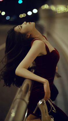 Woman in a red dress leaning on a railing at night with city lights in the background
