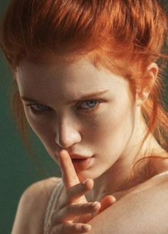 Woman with red hair and blue eyes, finger on lips, looking intensely at the viewer