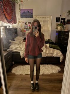 Person in a red sweater and denim shorts taking a mirror selfie in a bedroom