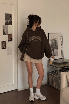 Woman in a brown hoodie and pleated skirt holding an iced coffee indoors