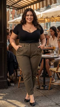 Curvy woman in a black top and olive pants smiling at an outdoor cafe