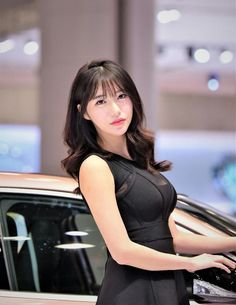 Woman in a black dress with long hair standing next to a car indoors