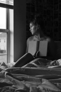 Woman with a messy bun holding a book in bed by a window