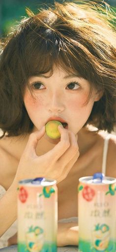 Woman with short hair and blush eating a fruit, with beverage cans in the foreground