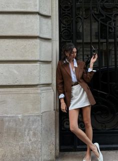Woman in a brown blazer and white mini skirt leaning against a stone wall