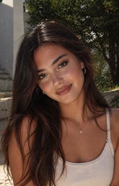 Woman with long dark hair wearing a white tank top and gold jewelry outdoors
