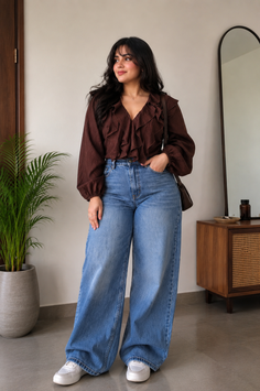 Woman with long dark hair wearing a brown ruffled blouse and wide leg jeans indoors
