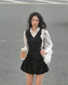 Woman with long curly hair in a black vest and skirt holding coffee on a street