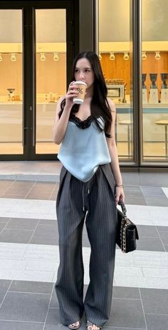 Woman with long black hair in a light blue camisole and pinstripe pants holding a coffee cup
