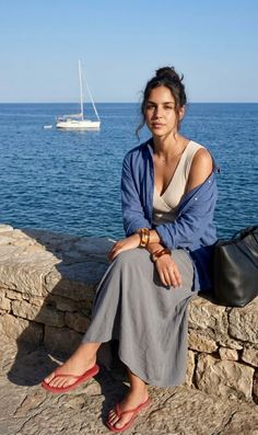Woman in a blue shirt and long skirt sitting by the sea with a sailboat in the background