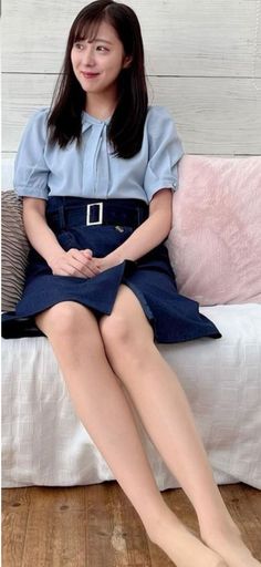Woman with long dark hair wearing a blue blouse and denim skirt, sitting indoors