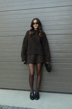 Woman in a dark sweater and mini skirt with sunglasses leaning against a wall