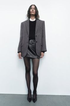 Woman in a gray blazer, black mini skirt, and black tights against a white wall