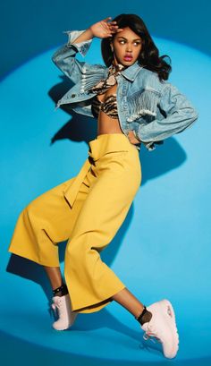 Woman in a denim jacket and yellow pants posing against a blue background