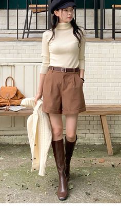 Woman in a cream turtleneck, brown shorts, and knee-high boots holding a cardigan
