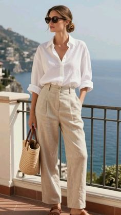 Woman in a white shirt and linen pants with a straw bag on a balcony overlooking the sea