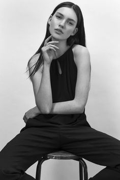 Woman with long dark hair in a black top and pants, sitting on a stool