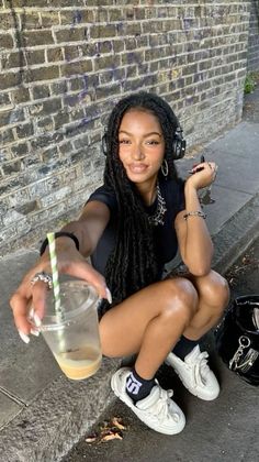 Woman with long dreadlocks and headphones squatting on a street, holding a drink